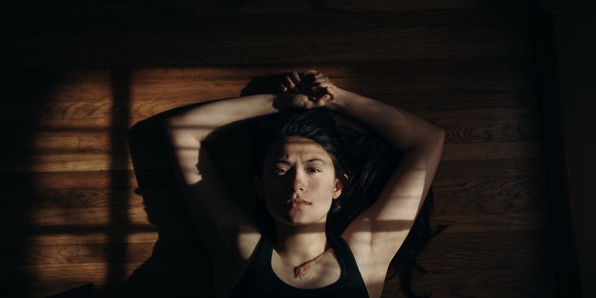 A color photograph from above of a white woman wearing a black tank top. She lays on a wood floor in the sunlight and shadow of a window, her arms above her head, looking into the camera.