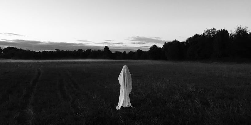 An eerie black-and-white photograph of a wide field with a figure dressed as a ghost at center.