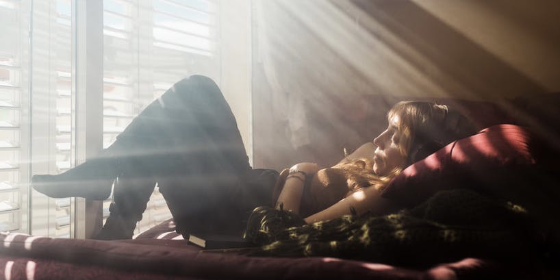 A color photograph of a window shining dappled light on the body of a young, thin white woman with blond hair wearing jeans and leaning back against pillows