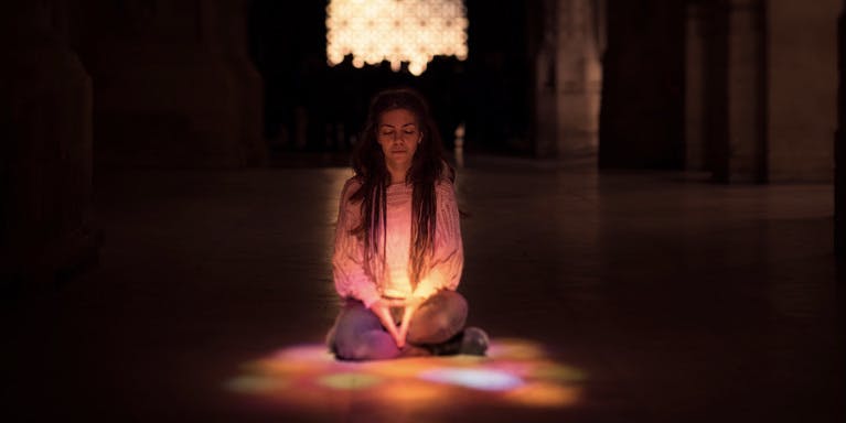 A color photograph of a young, thin white woman with long hair wearing jeans and a long-sleeve white shirt, sitting cross-legged and praying in the dappled color light of a church or temple.