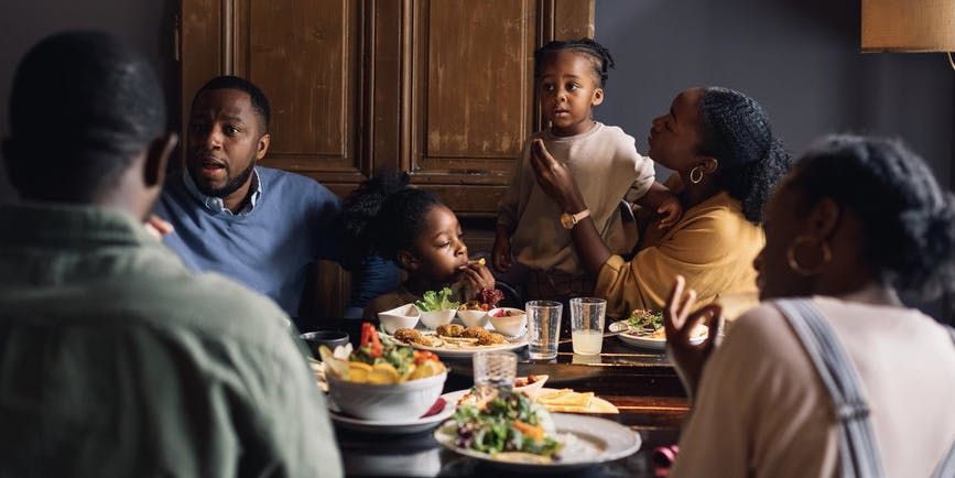A color photograph of a Black family gathered at their dining room table, with parents, children and teenagers mid-discussion.