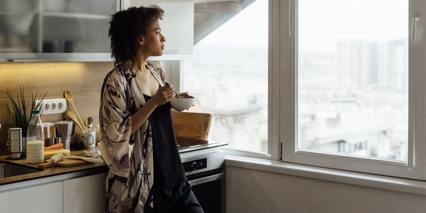 A color photograph of a young Black woman wearing a black jumper and colorful cardigan, standing in her brightly lit kitchen, eating from a bowl and looking out through wide windows.