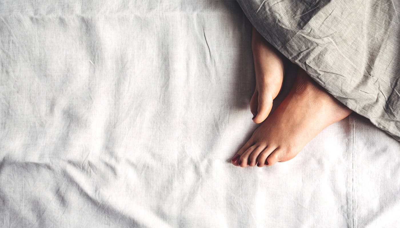 Dos pies que se asoman entre las suaves sábanas de lino, ambos apuntando hacia abajo