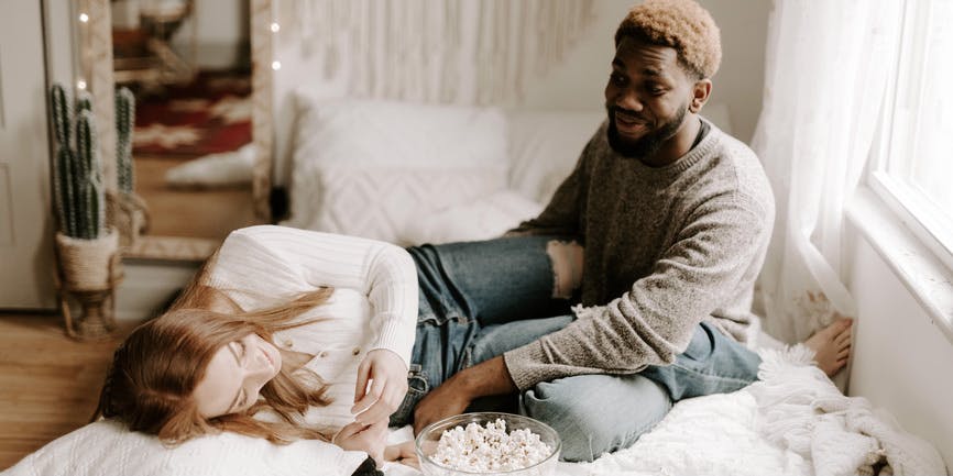 A color indoor photograph of a Black man with short brown hair wearing a long tan shirt and jeans and a white woman with long red hair wearing a long white shirt and jeans. They're on a bed with a bowl of popcorn, talking and laughing with each other.