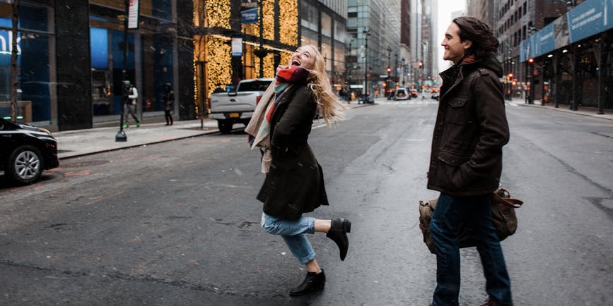 A color outdoor photograph of a young white woman and man in New York City during Christmas. They're wearing winter jackets and holding bags, smiling and laughing as they cross the street.
