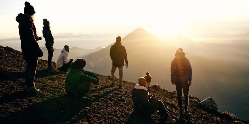 An outdoor color photograph of a mountain range. A group of young people in winter clothing stand and sit on one mountain top, looking away from the camera into the sunrise behind another mountain peak.