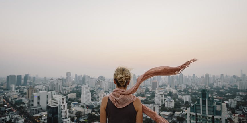 A color outdoor photograph of a white woman with blond hair tied up into a bun and a pink scarf flowing in the wind. She's standing with her back to the camera, with an expansive New York City scene and pink sky ahead of her.