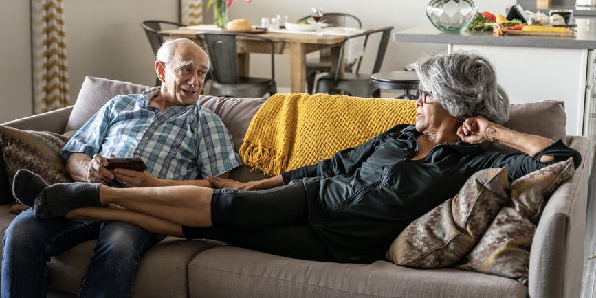 A color indoor photograph of a Hispanic senior couple on a couch. A gray-haired women wearing black stretches out with her feet on the lap of a grey-haired man wearing a blue plaid shirt and jeans, who rubs her feet.