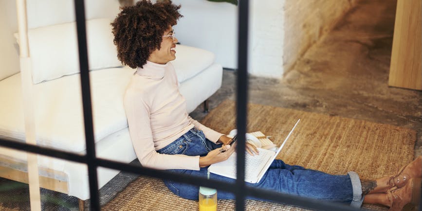 A color photograph taken through a window of a Black woman with an afro wearing a long sleeved shirt and jeans sits on the floor leaning against a couch, with a computer on her lap, smiling to someone off camera.