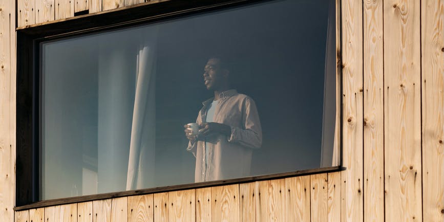 A color outdoor photograph taken from outside through a window, where a young Black man wearing a button down shirt stands holding a mug and gazing out.