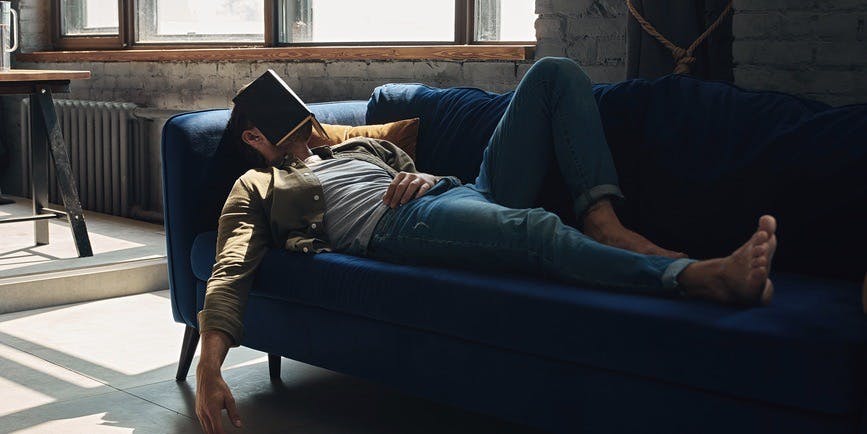 A color indoor photograph of a white man wearing jeans and a long-sleeved shirt, laying on a blue velvet couch with a book over his head, napping.