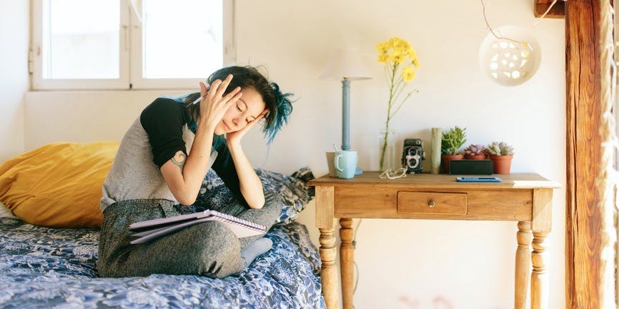 A color indoor photograph of a young Asian woman with long blue hair sitting on a bed with a blue coverlet and gold pillow, with a side table covered in flowers, a coffee cup, plants, etc. She's sitting on the bed wearing sweatpants and a long sleeve shirt with a notebook on her lap, her hands holding her head.