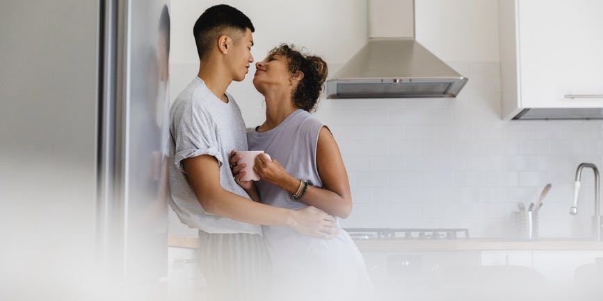 A color indoor photograph of a couple in a white and steel kitchen. A young Asian man with short hair wearing a gray t-shirt embraces a young brown-skinned woman, also wearing a short-sleeve gray tea, holding a coffee cup.