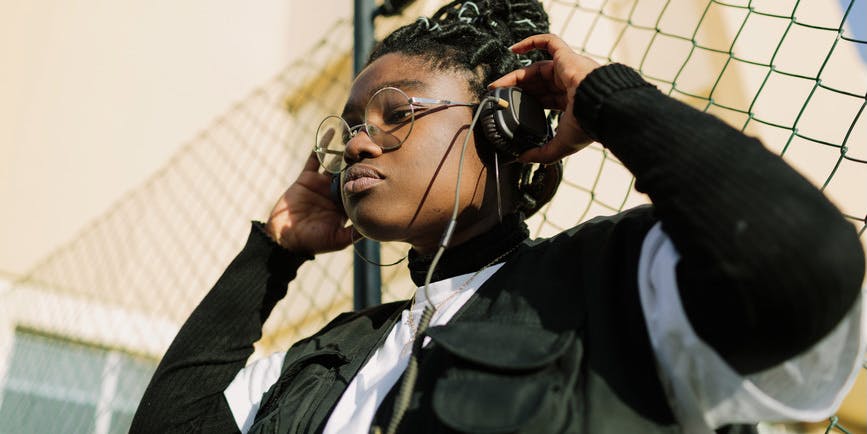 A color outdoor photograph of a young Black girl standing next to a fence. She's wearing a black and white jacket and holding headphones up to her ears.