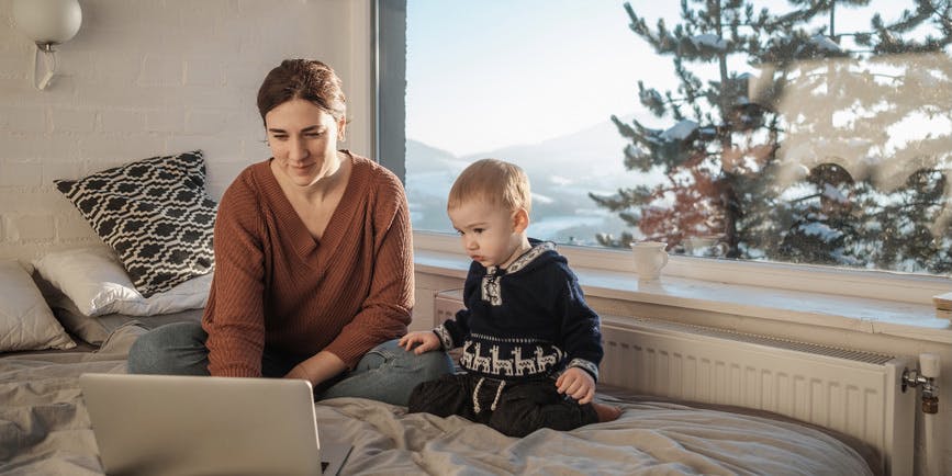 A color indoor photograph of a white mother wearing a brown sweater and her toddler, sitting on a bed in front of a computer, with a snowy tree-scape through the window behind them.