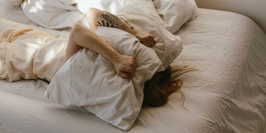 A white woman is lying on her back in bed with a pillow over her head, signifying discomfort