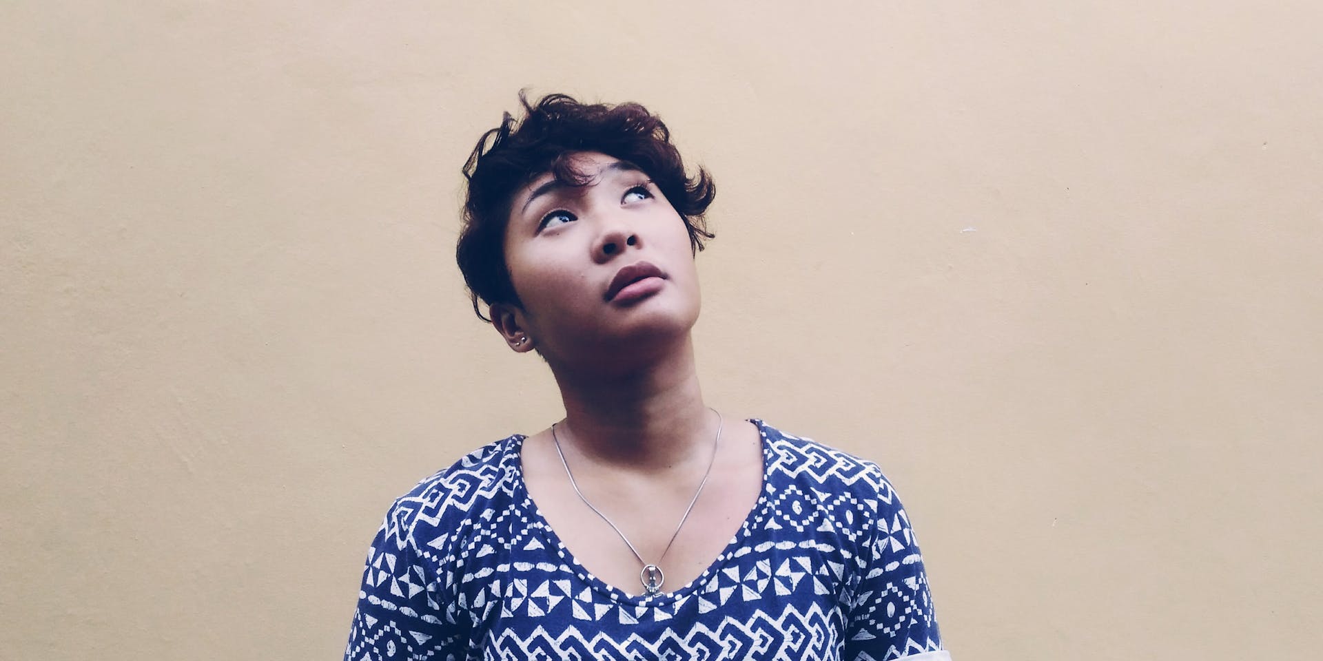 Una mujer filipina de pelo corto y oscuro está de pie contra una pared de color marrón claro, mirando hacia arriba, pensativa.