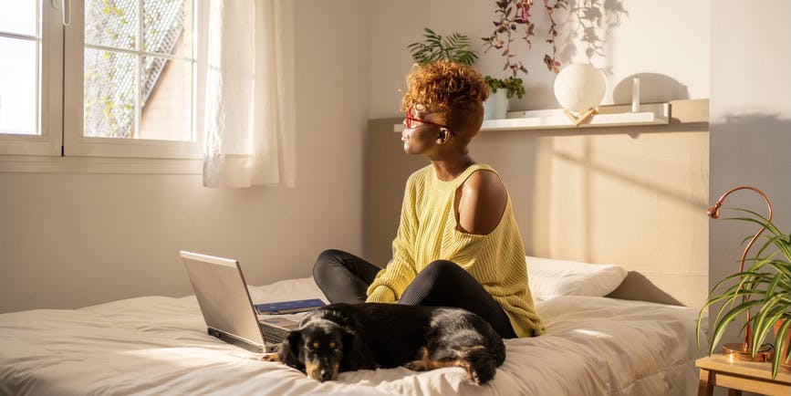 A color indoor photograph of a young Black woman with brown hair wearing a yellow sweater and black pants. They sit cross-legged on a bed in front of a laptop with a dog sleeping alongside them, staring out into the sunny window.