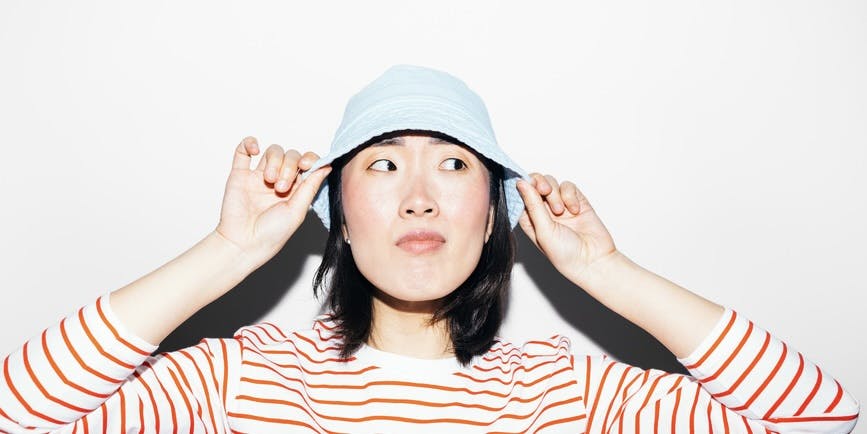 Asian woman with short dark hair and a striped red shirt holds a light blue bucket hat on her head while looking quizzically to the side