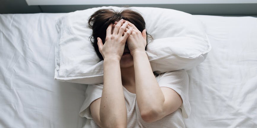 a white woman in a white t-shirt lays in bed on white sheets with her hands over her face in discomfort