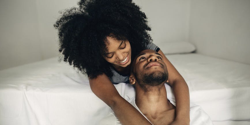 una joven negra con un afro está acostada en el borde de una cama abrazando a un hombre negro que está sentado en el suelo mirándola fijamente