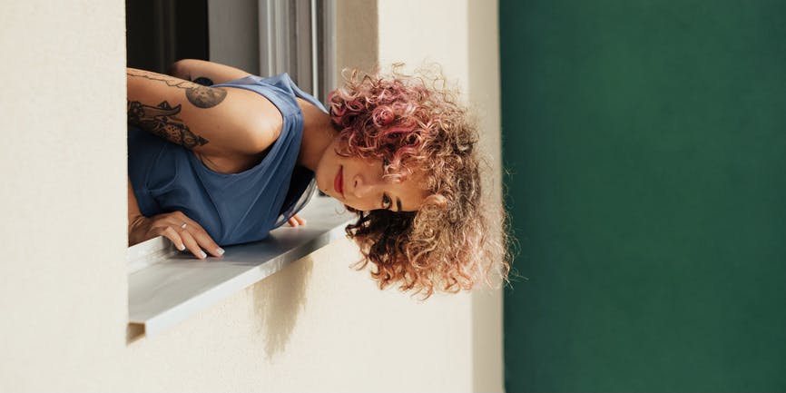 A tan-skinned woman with curly brown hair wearing a blue tank top with a few tattoos on her arm leans out of a window against a green wall. 
