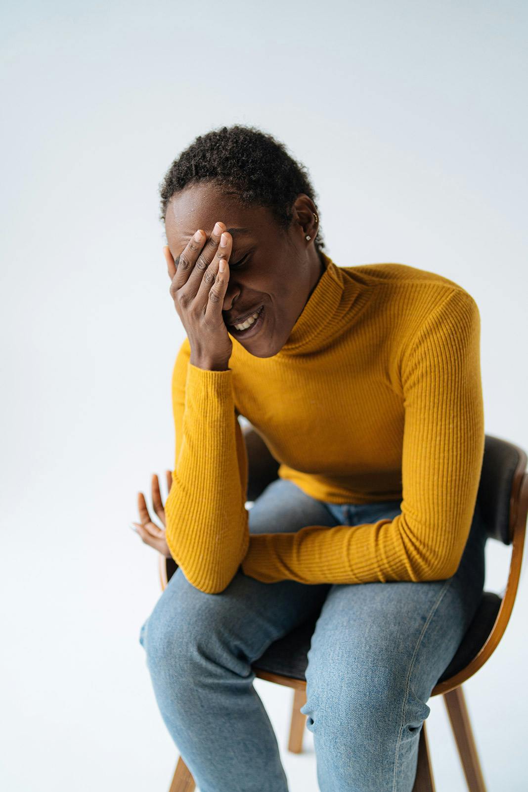 Young african american woman with short curly hair puts hand over her head and eyes, experiencing a migraine, sitting in a chair on a white background in the studio. Woman wearing a bright yellow turtleneck and blue jeans