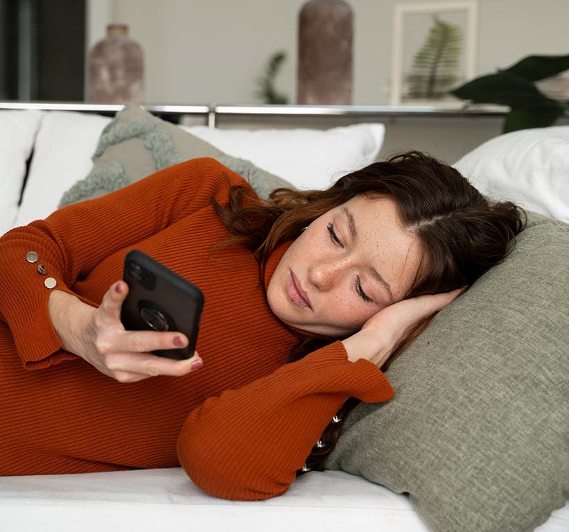mujer joven con cabello largo y castaño y vestida con una camisa roja, acostada de lado en el sofá, mirando su teléfono, que tiene en la mano