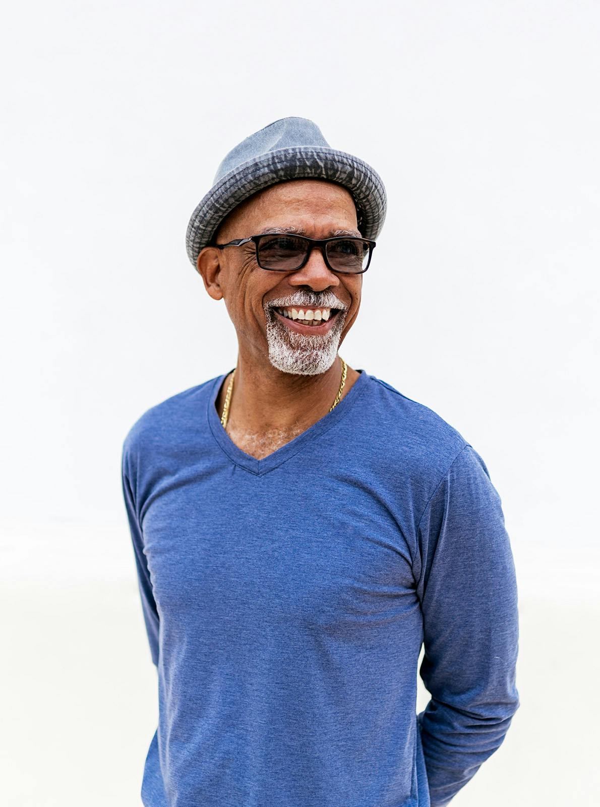 Un hombre negro mayor de pie frente a una pared blanca. Está sonriendo. Lleva gafas y sombrero.
