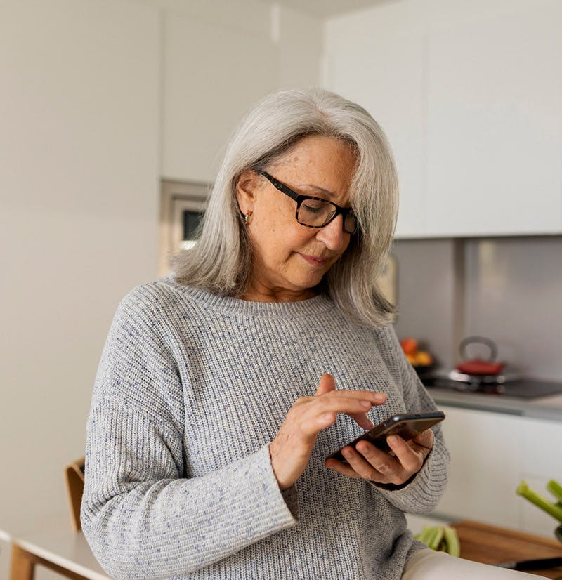 Mujer mayor usando un teléfono inteligente de pie en la cocina durante la mañana
