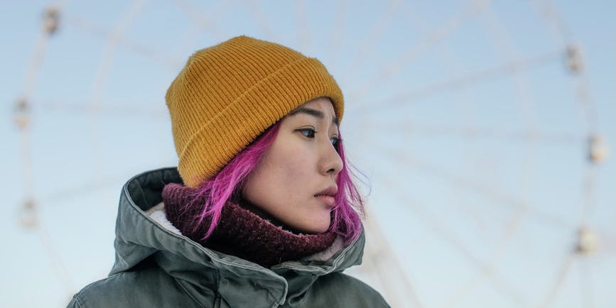 Portrait of a young Asian woman with pink hair wearing a yellow winter cap, thick coat and turtleneck sweater, looking off camera with the London Eye in the background.