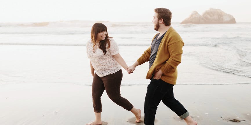 A curvy white man and woman wearing pants and long shirts walk barefoot on a beach.