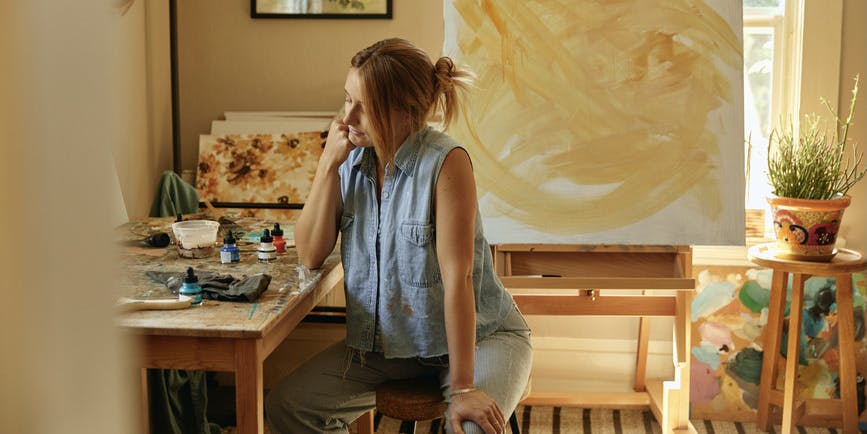 An indoor photograph of a white, thin woman with blond hair tied up into a knot wearing a paint-covered denim tank and carbo pants. She's sitting at a desk in a painting studio, resting her face on her hand, looking downward.