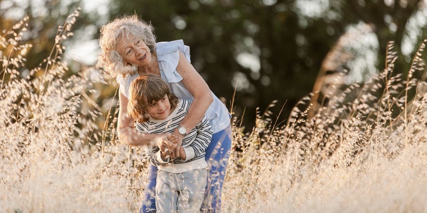 A color outdoor photograph of an older white woman with gray-blond hair, wearing jeans and a blue-striped shirt, standing in a field of wild grass and hugging her grandchild, a white boy with long blond hard wearing gray jeans and a striped shirt.