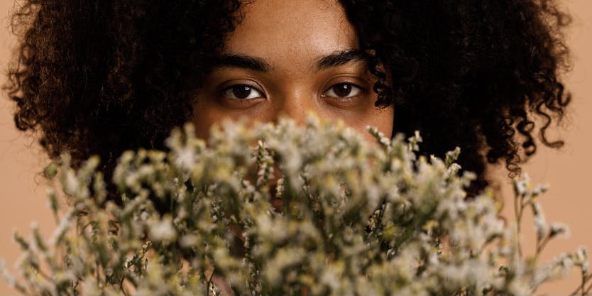 A closeup photograph of a Black woman with curly brown hair standing in front of a peach-colored wall. She's holding a bouquet of wildflowers close to her face, so only her swollen eyes are visible behind it.