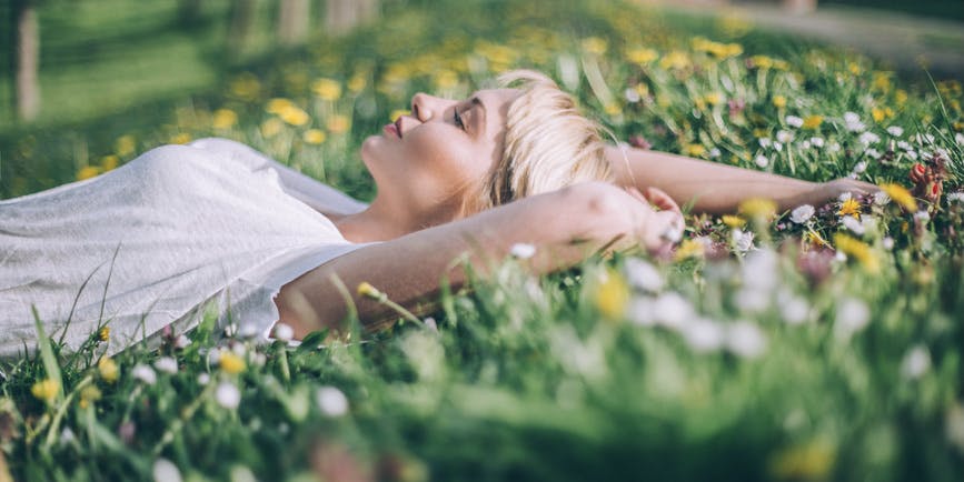 A color photograph of a young white woman with short blond hair wearing a white t-shirt, laying outside in a field of wildflowers with her face up to the sky.