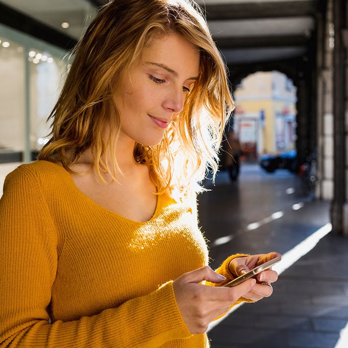 Mujer joven con un suéter amarillo naranja en su teléfono, afuera, sonriendo sutilmente mientras mira el teléfono