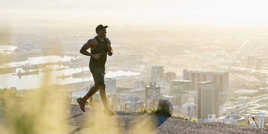 A fit Black man road running at sunrise. He is well built and wears all black sportswear whilst running on a road overlooking a city at sunrise.