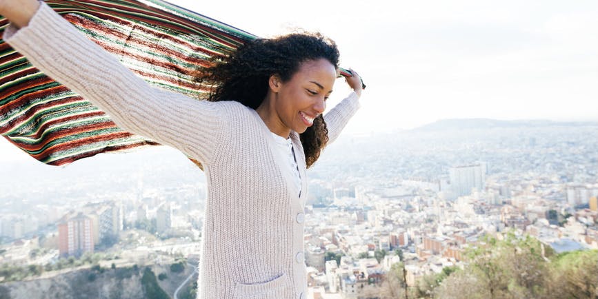 A young Black woman wearing a white sweater smiles and holds up a blanket behind her on a windy day with a cityscape in the distance behind her.
