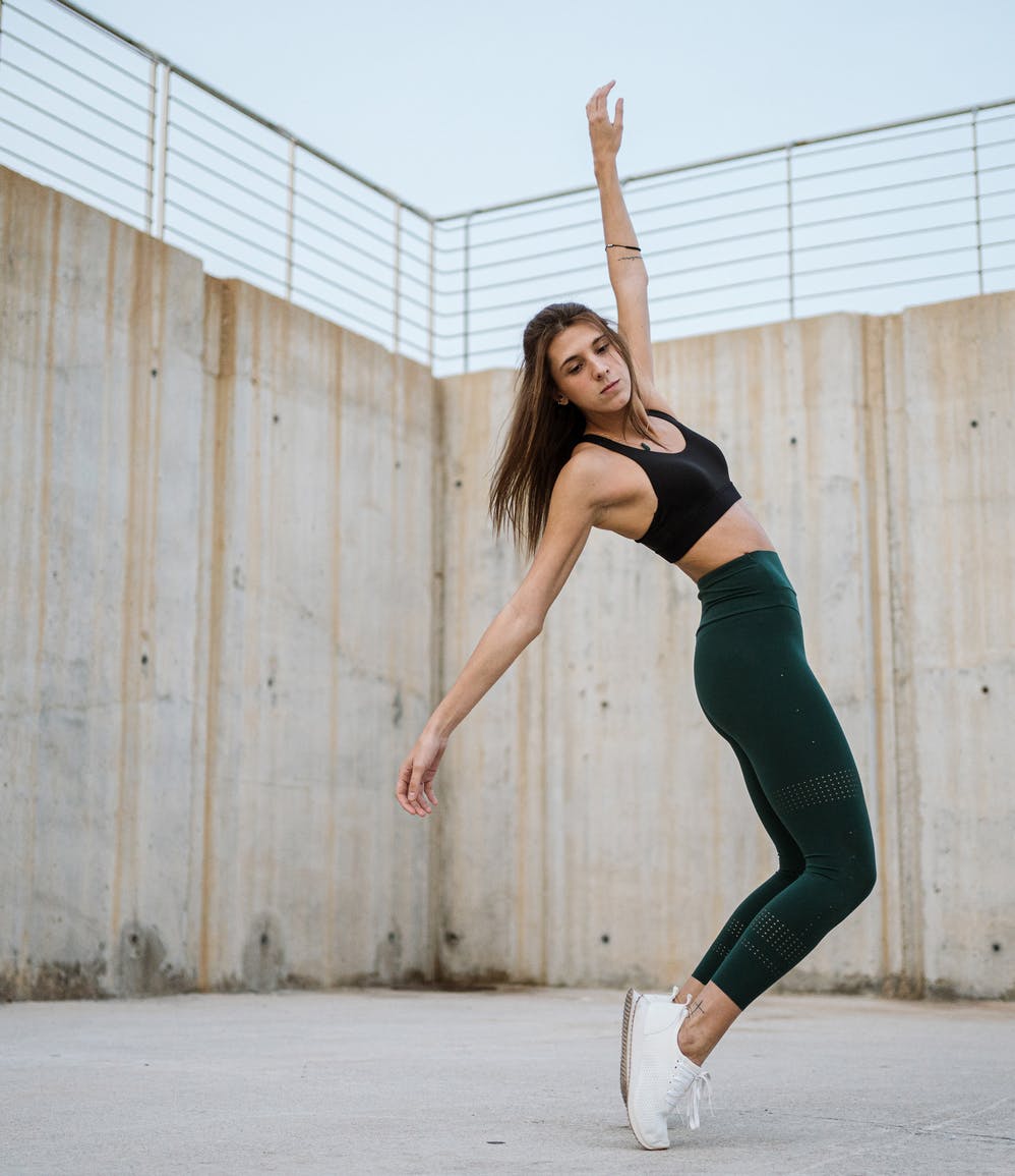 Vista lateral de una joven en ropa deportiva balanceándose de puntillas con los brazos extendidos mientras baila fuera de un edificio de concreto erosionado en la ciudad