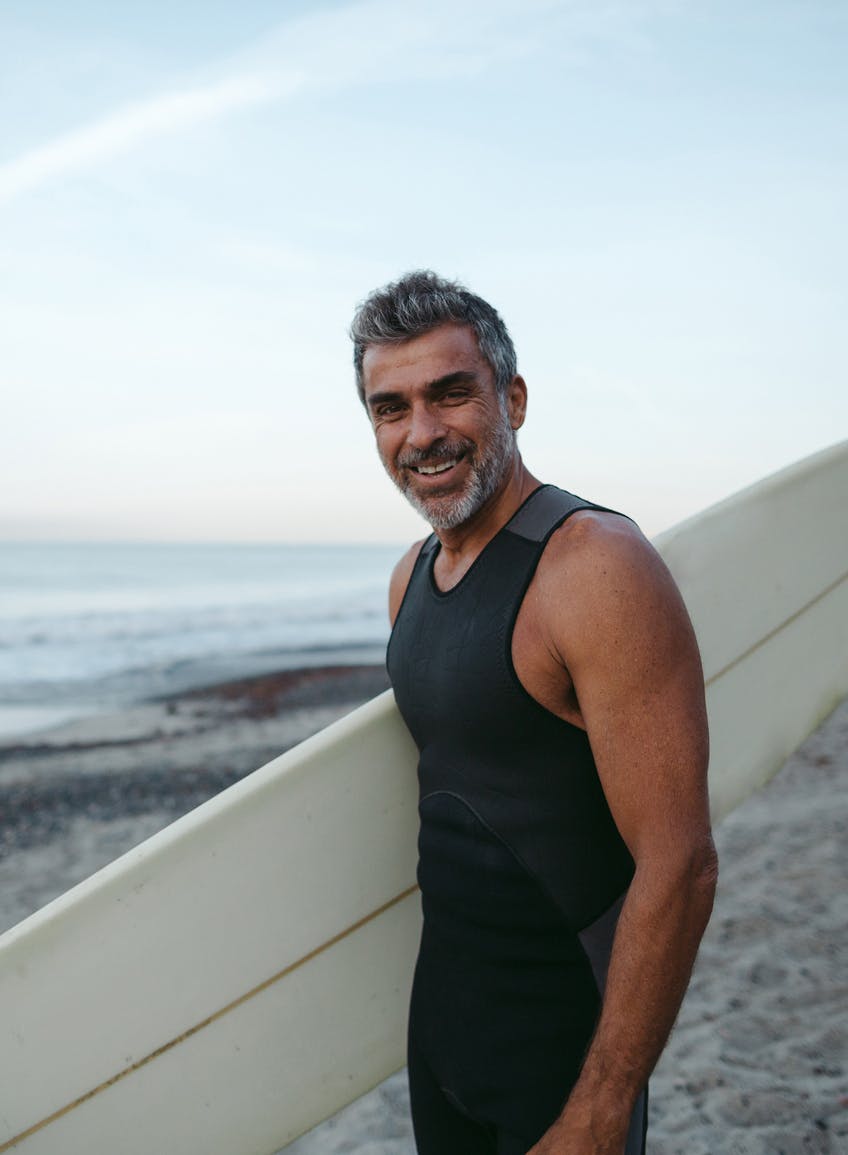 A smiling middle-aged man with greying hair in a wetsuit carrying a surfboard on the beach