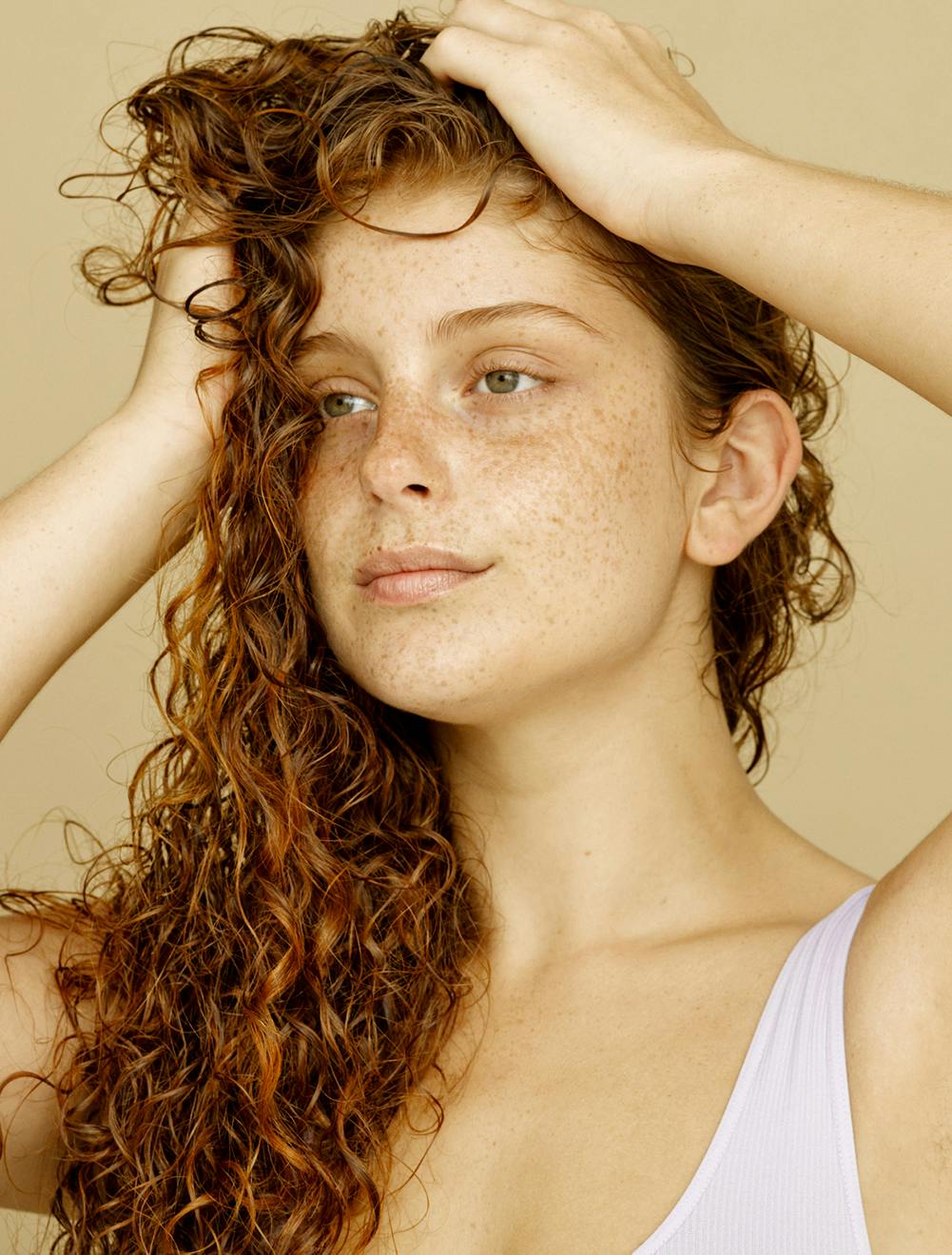 Woman with curly red hair and stark freckles holding up her hair with both hands, looking into the distance