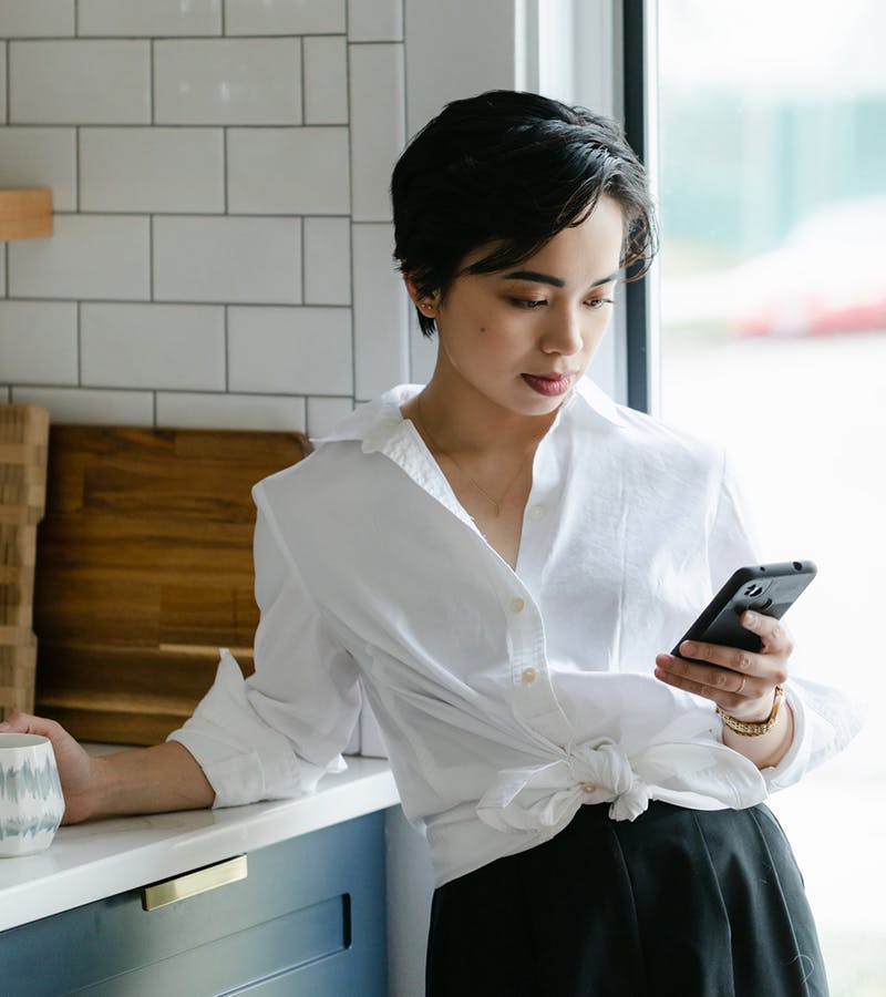 Young Asian woman standing in a kitchen holding a mug and looking at her phone