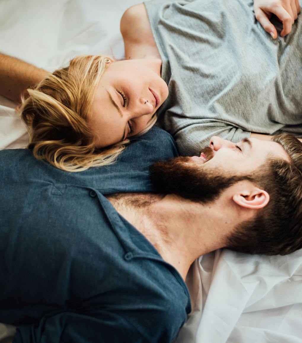 A young couple – a woman with blonde hair and a bearded man with brown hair – with eyes closed on a bed snuggling.