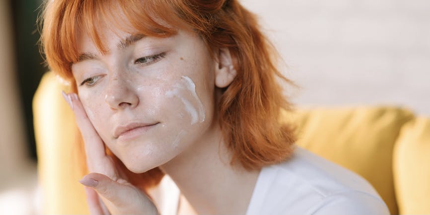A young white woman with fair skin and short red hair wearing a white t-shirt puts moisturizer on her face while looking off camera.