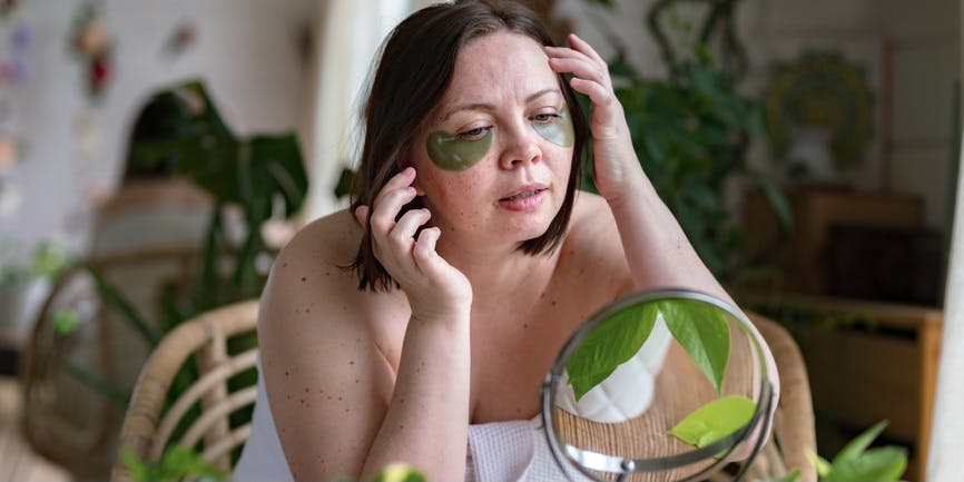 A curvy white woman with short brown hair wears a towel wrapped around her. She’s in a room full of plants, looking into a tabletop mirror while applying eye masks below her eyes.