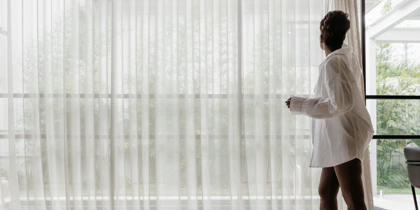 A Black woman with brown hair wearing a long white shirt opens the sheer blinds covering a large window.