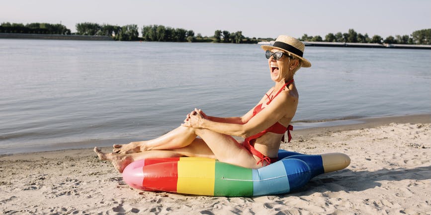 An older thin white woman with short white hair wearing a sunhat, sun glasses and a bathing suite sits on a rainbow colored raft on the sand in front of the water.