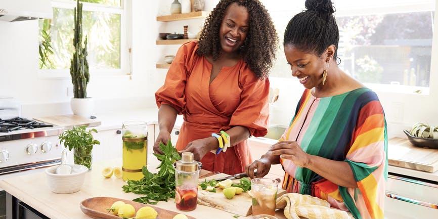 Two young and beautiful Black woman wearing dresses stand at a kitchen counter making cocktails with fresh fruit and herbs.