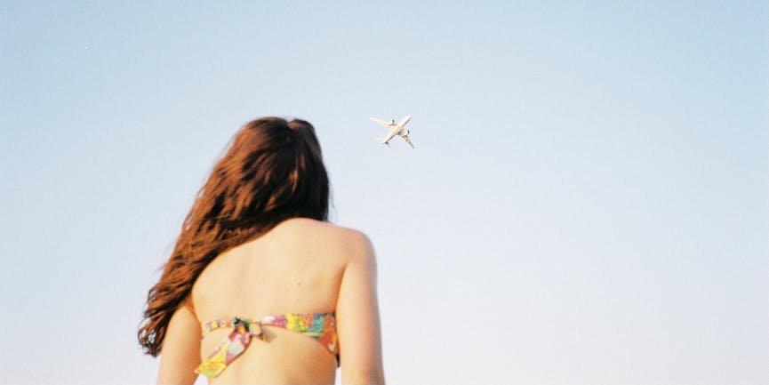 A young white girl wearing a rainbow bikini top looks away from the camera and up into the sky where a plane flies.